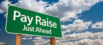 Green road sign with 'pay raise just ahead' text against a blue sky with clouds.